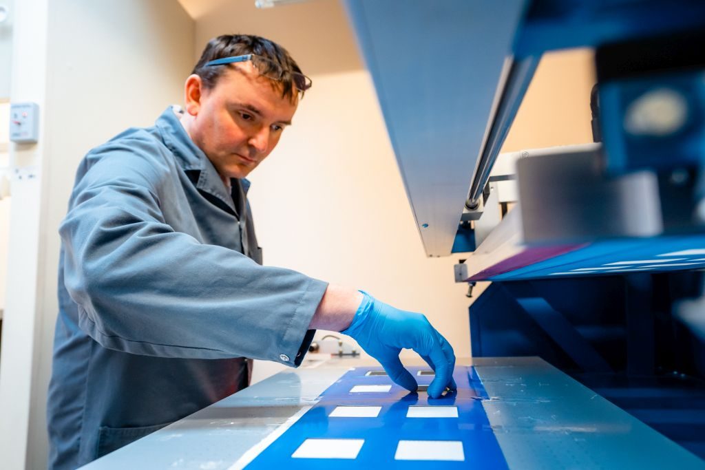 Zdjęcie przedstawia mężczyznę ubranego w niebieski fartuch i rękawiczki, który układa elementy do drukowania warstwy katodowej