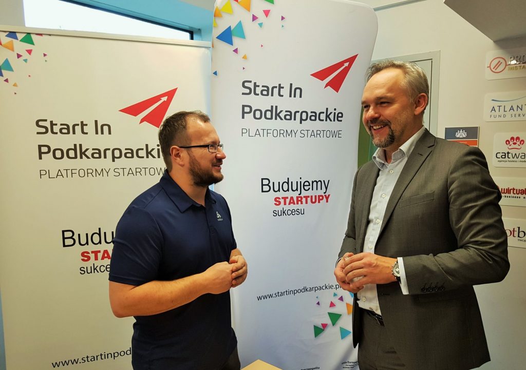 Zdjęcie przedstawia dwóch mężczyzn rozmawiających na tle planszy Platformy Startowej Start In Podkarpackie podczas spotkania informacyjno-promocyjnego w Kielcach.