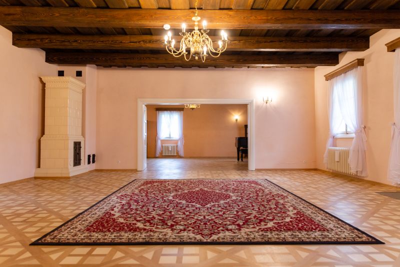 Reprezentacyjny salon z drewnianym, belkowanym sufitem, taflowym parkietem, na którym leży bordowy dywan. w lewym rogu pokoju biały kaflowy piec. Wnętrze oświetla żyrandol
