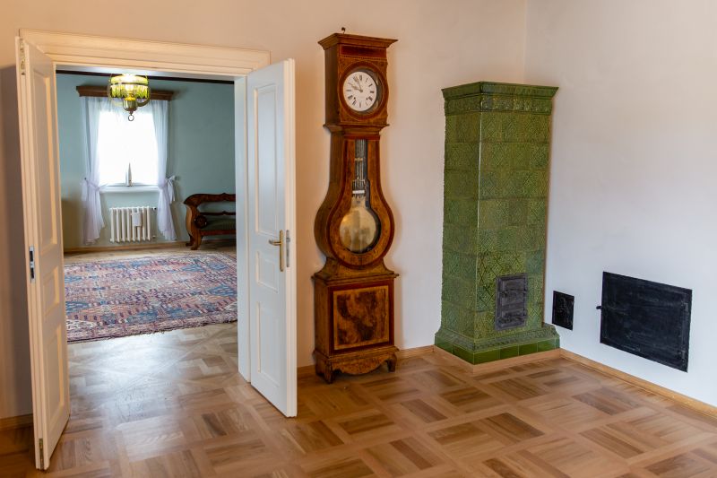Jedno z pomieszczeń, w którym w prawym rogu ustawiono piec z ciemnozielonych kafli. obok pieca stoi zabytkowy drewniany zegar. pośrodku otwarte drzwi do sąsiedniego pomieszczenia
