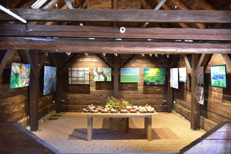 wnętrze zabytkowej, drewnianej stodoły z Cieszanowa. Na pierwszym planie stół w owocami, w tle powieszone na ścianach kolorowe brazy