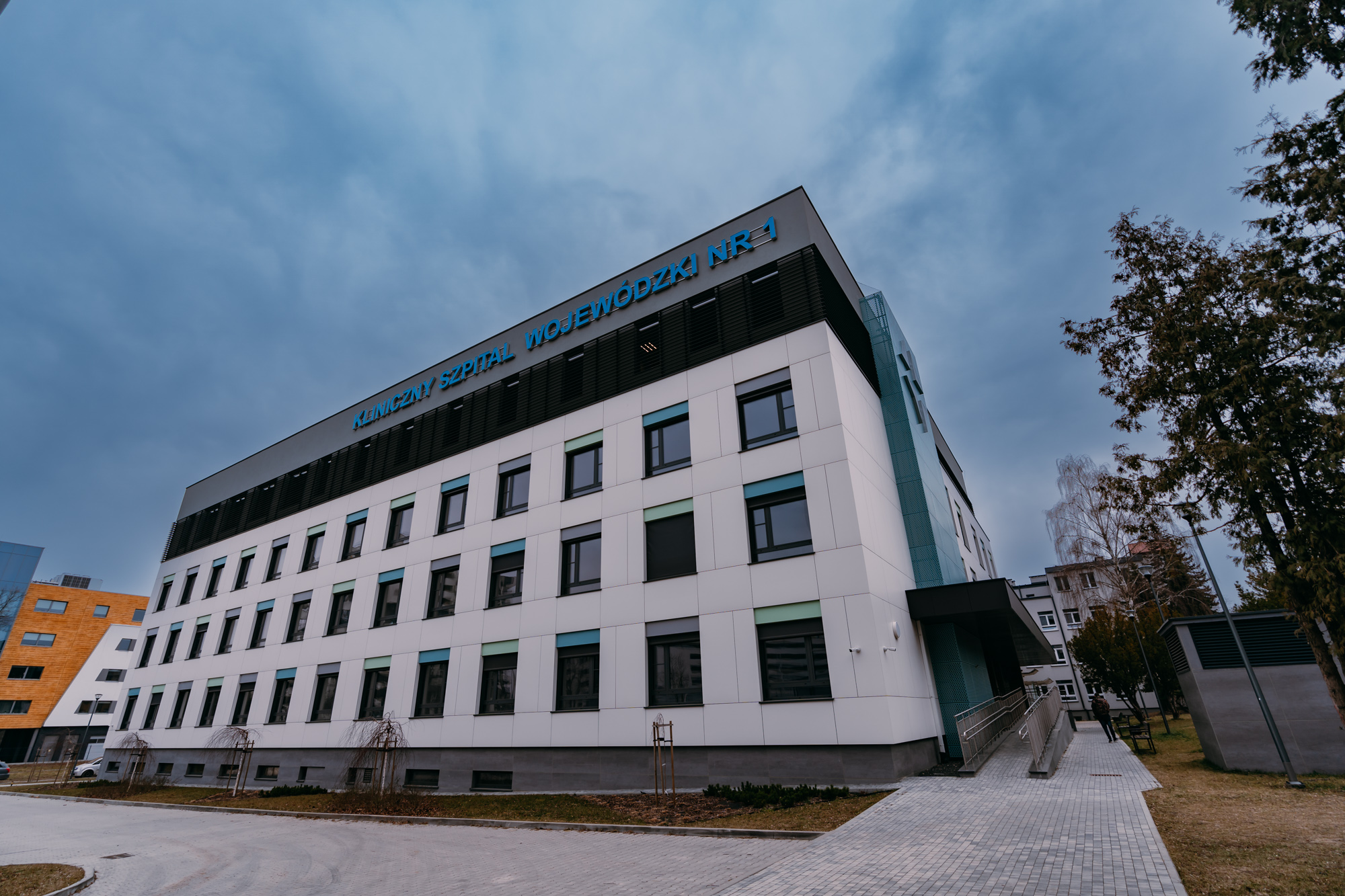Biała fasada budynku, na górze budynku, na szarym tle niebieskie litery tworzące napis: Kliniczny Szpital Wojewódzki nr 1