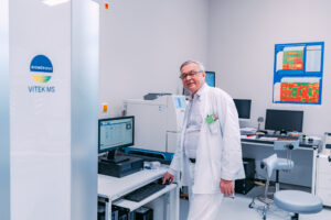 W fartuchu, ubrany na biało, starszy, siwy mężczyzna w okularach – dr Wojciech Kądziołka stoi w laboratorium przy biurku z komputerem