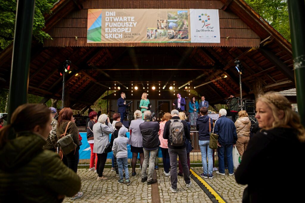 Osoby stojące przed sceną widoczne tyłem. Na drewnianym zadaszeniu widoczny baner z napisem: Dni Otwarte Funduszy Europejskich i logotypem DOFE.