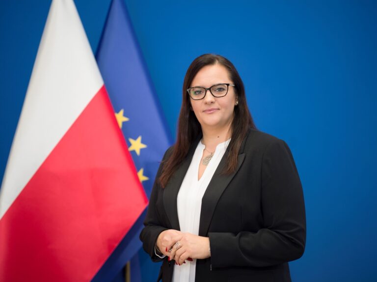 Małgorzata Jarosińska-Jedynak, sekretarz stanu w Ministerstwie Funduszy i Polityki Regionalnej. Brunetka w okularach, ubrana w czarną marynarkę i białą bluzkę stoi na niebieskim tle, po lewej biało-czerwona flaga Polski i flaga UE: niebieska w żółte gwiazdki