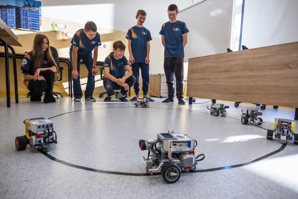 Grupa uczniów na zajęciach z robotyki. Obserwują roboty poruszające się po podłodze.