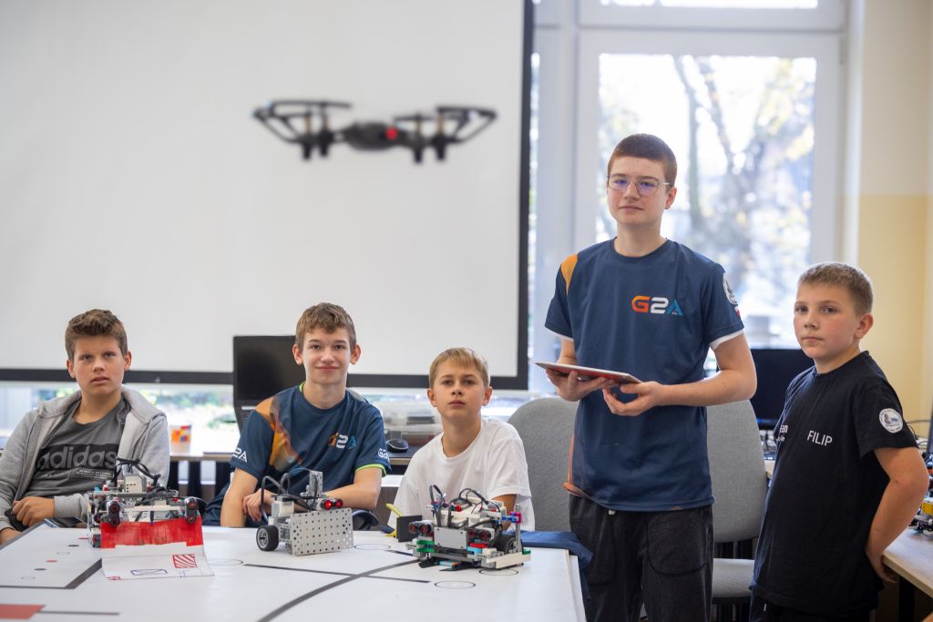 Grupa uczniów uczestniczy w zajęciach z robotyki. Część z nich siedzi wokół stołu z robotami LEGO. Jeden stoi obok stołu, trzyma tablet i nadzoruje drona unoszącego się w powietrzu. Wszyscy są ubrani w koszulki związane z robotyką.