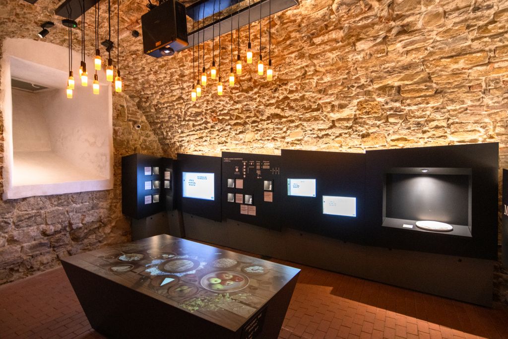 Jedno z pomieszczeń Podziemnej Trasy Turystycznej. Na środku wyeksponowany stół multimedialny. Surowe ściany z cegły i przyćmione oświetlenie tworzą klimat tego miejsca.