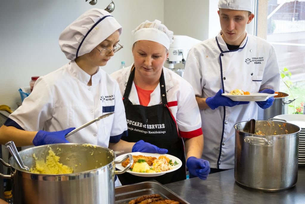 Dwóch uczniów w białych kucharskich uniformach przygotowuje się do rozdania posiłków. Jedna z nich, ubrana w rękawiczki ochronne, trzyma w rękach talerz z jedzeniem. Starsza kobieta i chłopak przyglądają się.