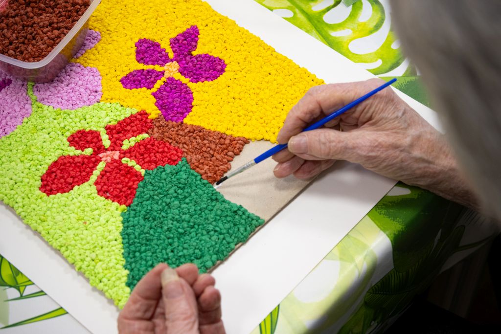 Zbliżenie na ręce osoby starszej, która wykonuje kolorową, kwiatową mozaikę z kawałków papieru.