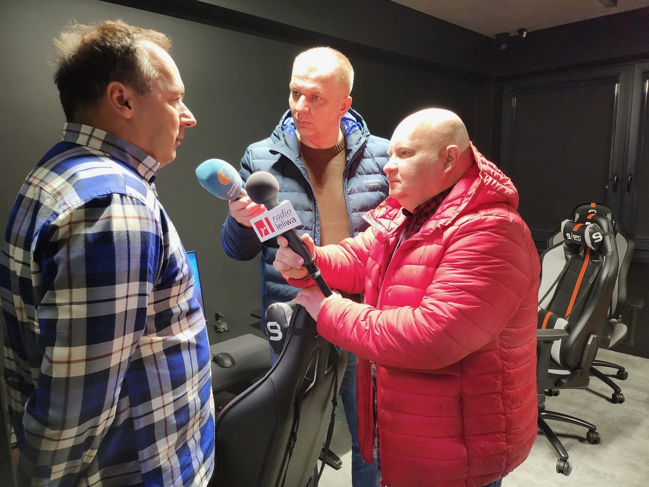 Wywiad radiowy w trakcie nagrywania. Dwaj mężczyźni , jeden w czerwonej, drugi w niebieskiej kurtce przeprowadzają wywiad radiowy z innym mężczyzną w kraciastej koszuli.