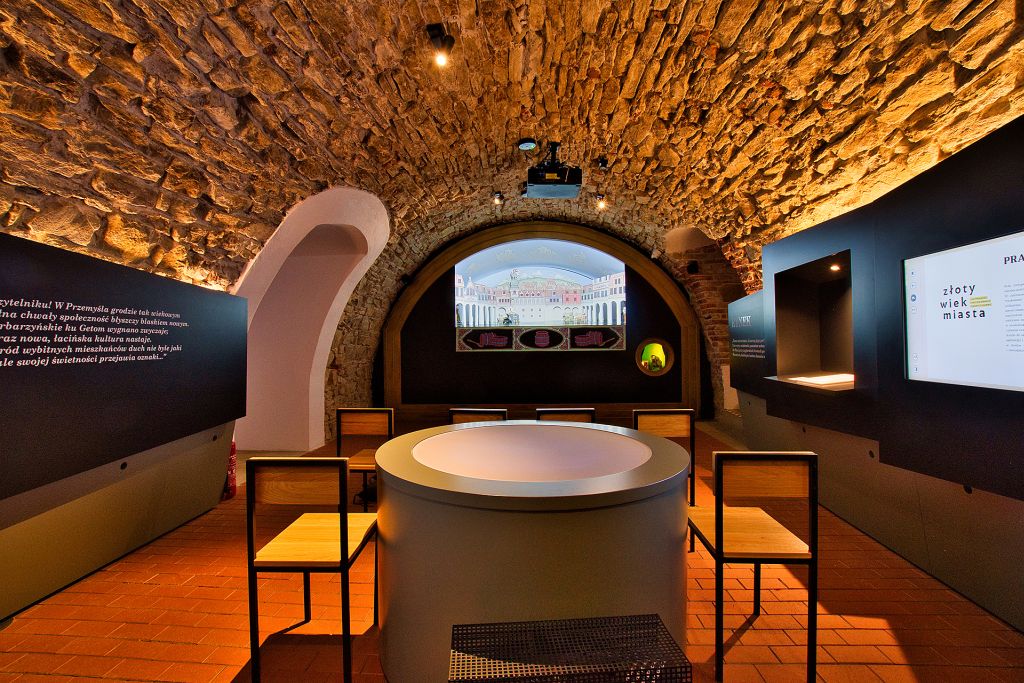 Pomieszczenie w podziemiach muzeum. Ściany z surowego kamienia. Widoczne ekspozycje muzealne i ekrany na ścianach. Po środku okrągły stów i dwa krzesła.