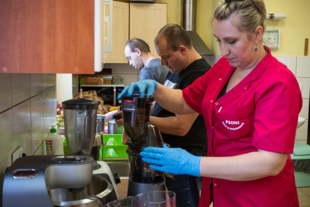 Trzy osoby pracują w kuchni i przygotowują jedzenie. Kobieta w czerwonym uniformie i niebieskich rękawiczkach obsługuje blender. Na blacie jest dużo kuchennych przyrządów i sprzętów.