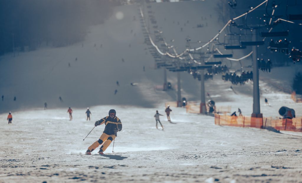 Narciarz dynamicznie zjeżdża po stoku narciarskim. W tle liczni narciarze i wyciąg krzesełkowy