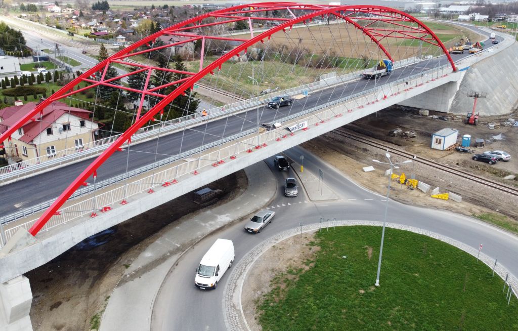 Na pierwszym planie okazały most w kolorze czerwonym. Pod mostem rondo, po którym jadą cztery samochody. W tle widoczna budowa i panorama miasta.